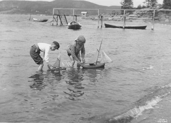 Gutter leker med seilbåter på Sundestranda, badebryggen i bakgrunnen, lengre bak et gammelt badehus (Wilse, 1904)